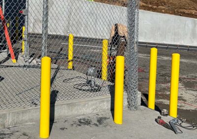 A row of bright yellow bollards lines the edge of a concrete platform near a chain-link fence, with construction tools and equipment scattered around in an outdoor work area.