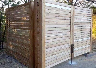 A modern wooden fence enclosure with horizontal slats, featuring a double-door gate with vertical handles, set outdoors on a concrete base surrounded by trees and foliage.
