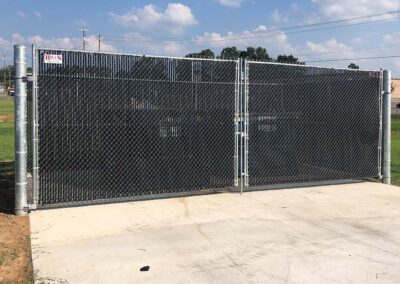 A double chain-link gate with privacy slats stands closed on a concrete pad, surrounded by grass and trees under a partly cloudy sky.