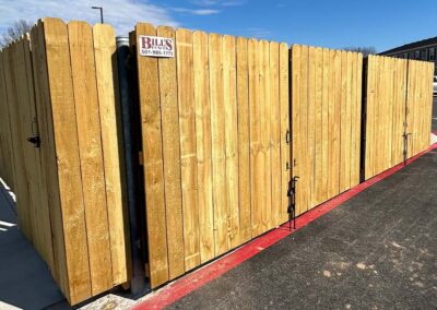 A large wooden fenced enclosure with a gate sits on a paved surface next to a red-painted curb, under a clear blue sky. A sign with contact information is attached to the fence.