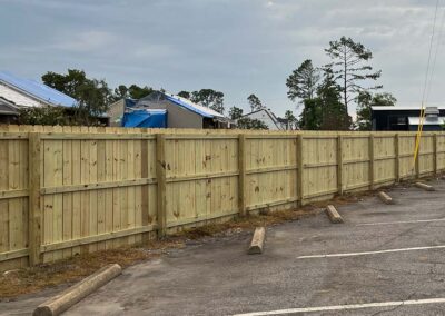 A light wooden privacy fence lines the edge of a parking lot with worn asphalt and empty concrete parking stops. In the background, damaged buildings with blue tarps are visible under a cloudy sky.
