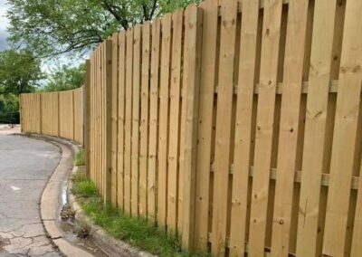 A new wooden fence runs along the edge of a curved sidewalk and street, with green grass and trees visible behind it under a cloudy sky.
