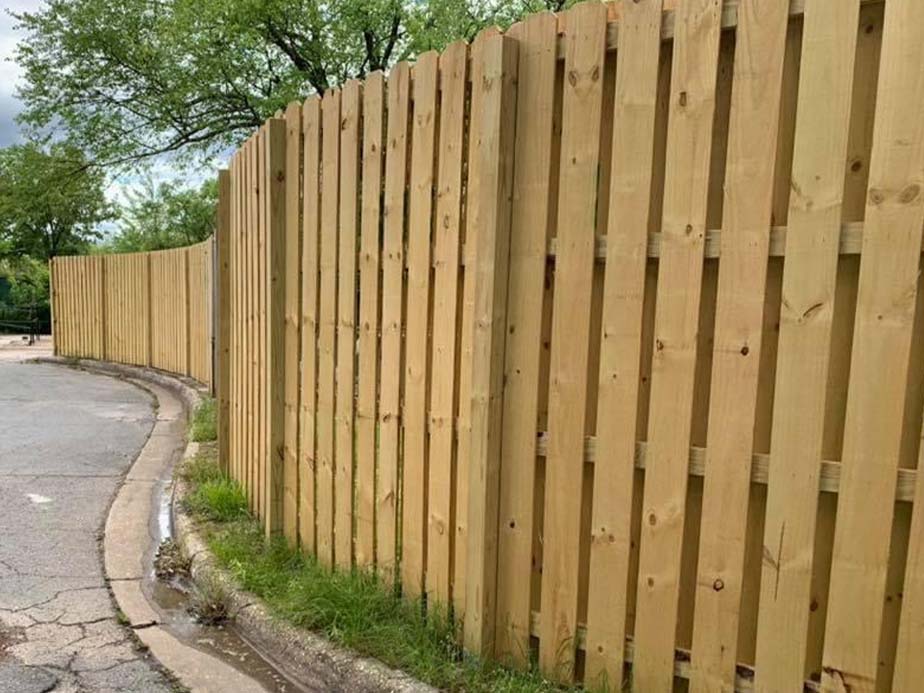 A new wooden fence runs along the edge of a curved sidewalk and street, with green grass and trees visible behind it under a cloudy sky.