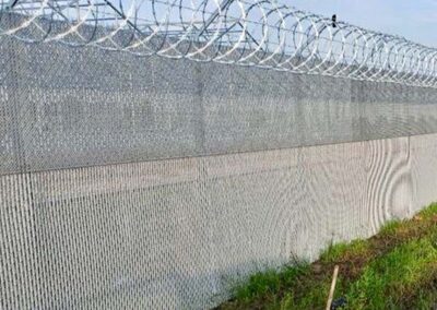 A tall metal fence topped with rolls of razor wire stands beside a grassy area. The fence casts shadows on itself, and the sky in the background is clear and blue.