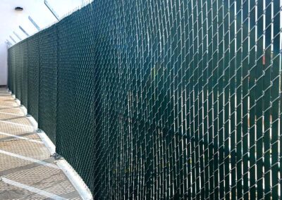 A tall chain-link fence with green privacy slats runs alongside a concrete pathway, topped with angled security arms and barbed wire. Shadows from the fence create a striped pattern on the ground.