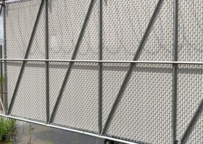A large metal gate with a chain-link pattern, reinforced with diagonal and horizontal bars, stands on wheels over a wet, slightly overgrown ground.