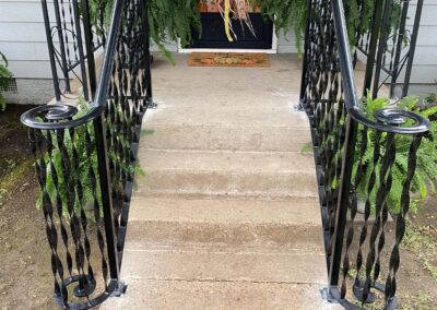 Concrete porch steps with black decorative metal railings on both sides, leading up to a door with a fall-themed doormat and green ferns placed on either side of the entryway.