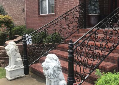 Two white lion statues guard the base of brick steps with ornate black wrought iron railings, leading to a red-brick house entrance. Greenery and a potted plant are visible nearby.