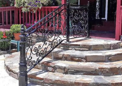 Curved stone steps lead up to a porch with a decorative black wrought iron railing featuring intricate scrollwork. The background includes red wooden railings, potted plants, and flowers.