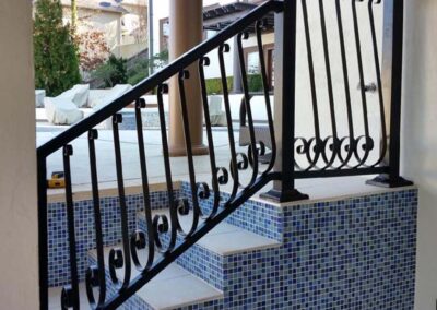 A black metal railing with ornate designs lines a staircase covered in small blue and white mosaic tiles, leading up to an outdoor patio area.