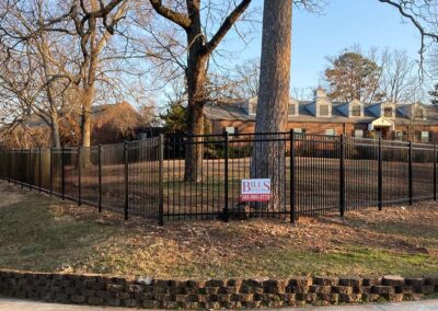 Black metal fence with vertical bars encloses a yard with leafless trees and a brick building in the background. A sign on the fence reads “BILL’S” with a phone number. The scene appears to be in late autumn or winter.