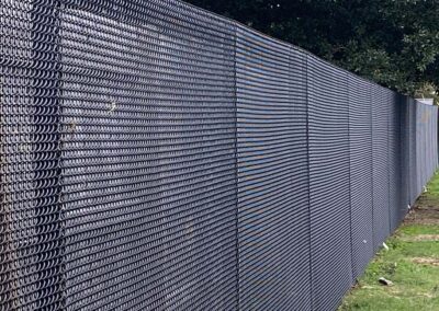 A long chain-link fence covered in black privacy slats runs alongside a grassy area, with trees visible in the background.