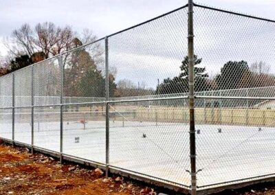A fenced concrete area, possibly a tennis or basketball court, surrounded by bare trees and dirt. The sky is overcast, and there are no people present.