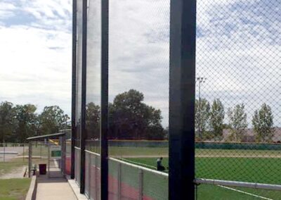 A tall protective net stretches along the edge of a baseball field, with green grass, trees, and a blue sky with scattered clouds in the background. A dugout and walkway are visible on the left.