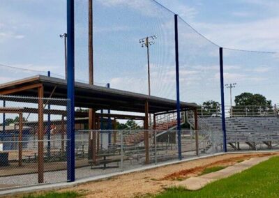 A covered seating area and metal bleachers are seen behind chain-link and netted fences at an outdoor baseball or softball field under a partly cloudy sky.