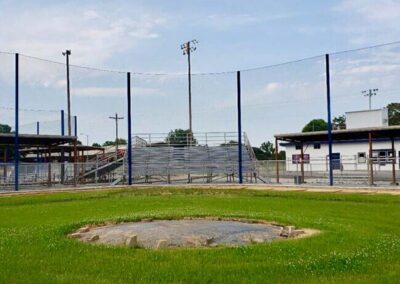 An empty baseball field with a worn pitchers mound and green grass, surrounded by a chain-link fence. Metal bleachers and a small building are visible in the background under a partly cloudy sky.