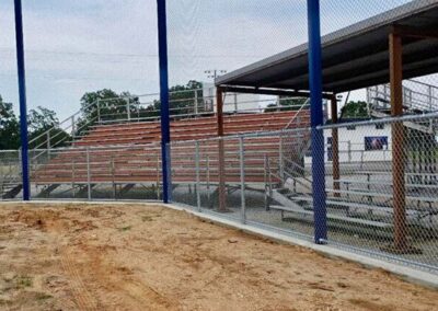 Bleachers behind a chain-link fence at a baseball or softball field, with some covered seating and dirt ground in the foreground. Trees are visible in the background under a partly cloudy sky.