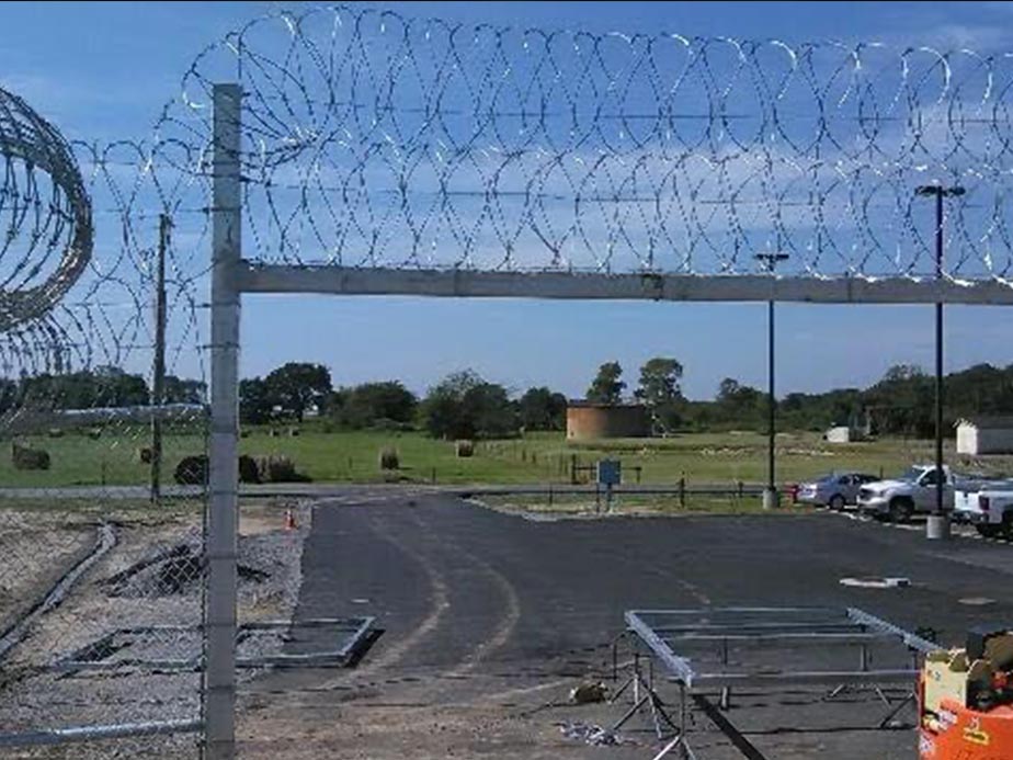 A fenced area with razor wire on top surrounds a paved lot with parked vehicles, construction materials, and equipment. Beyond the fence, there are green fields, trees, and a blue sky with scattered clouds.