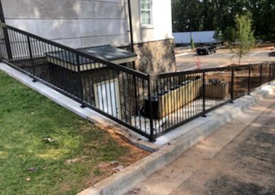 A black metal railing encloses a sloped walkway that leads down to a basement entrance at the side of a building. There is a grassy area on the left and a parking lot with trees in the background.