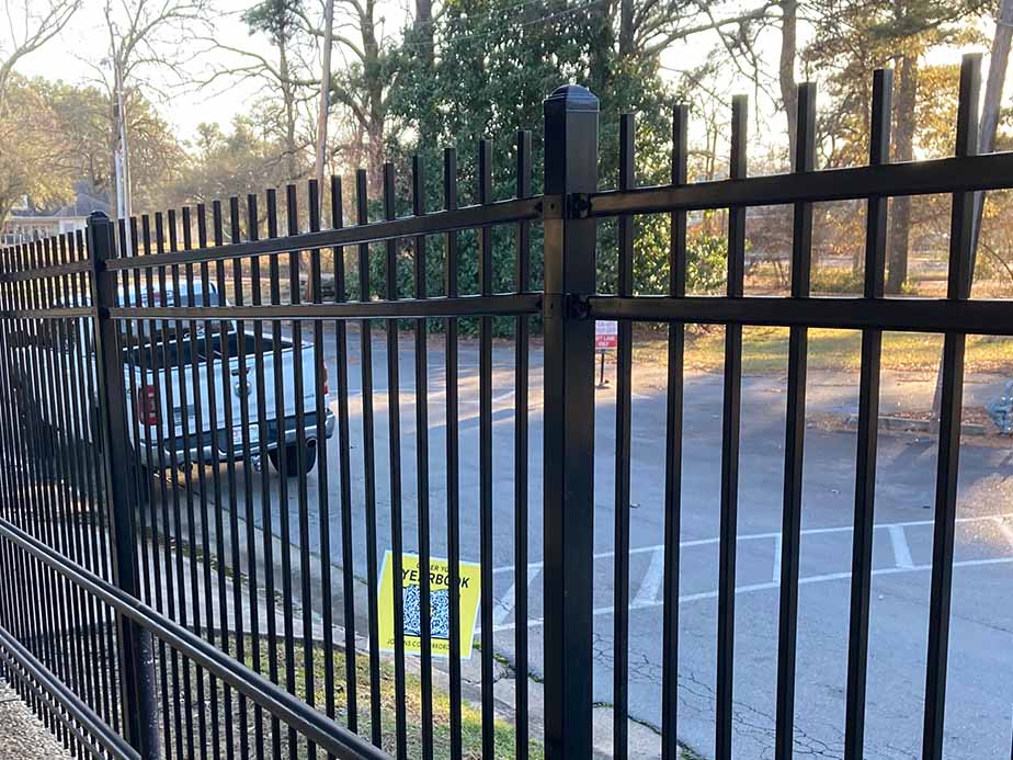 A black metal fence with vertical bars is in the foreground. Behind the fence, there is a white pickup truck parked in a lot, and a yellow sign is visible on the ground near the fence. Trees are in the background.