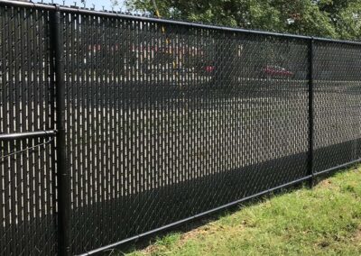 A black chain-link fence with privacy slats runs along the edge of a grassy area, with trees and a street visible in the background through the gaps at the top of the fence.