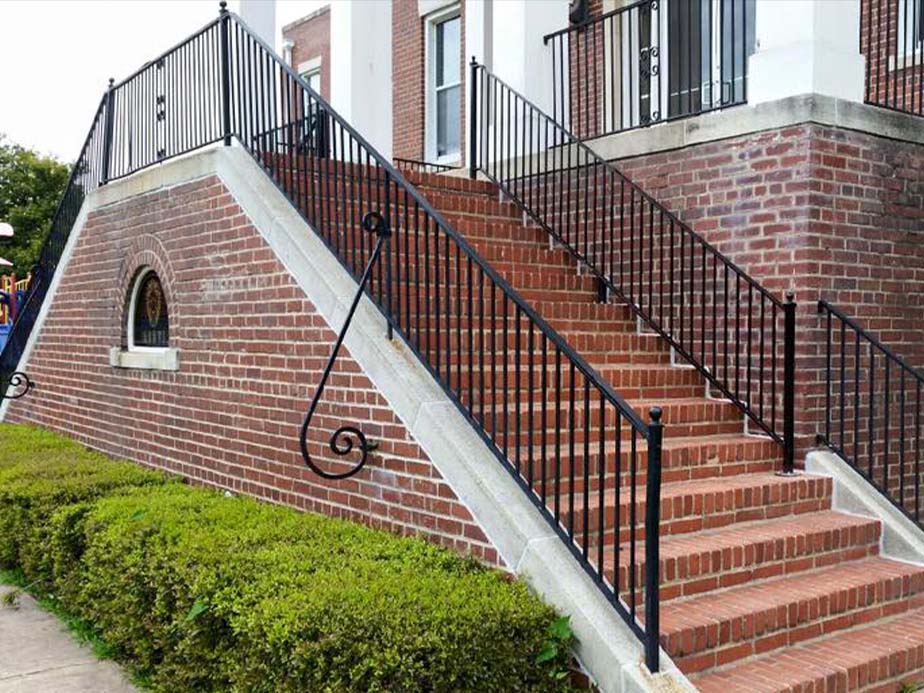 A brick staircase with black metal railings ascends alongside a brick building. Green hedges line the walkway at the base, and there is a small arched window set into the brick wall beneath the stairs.
