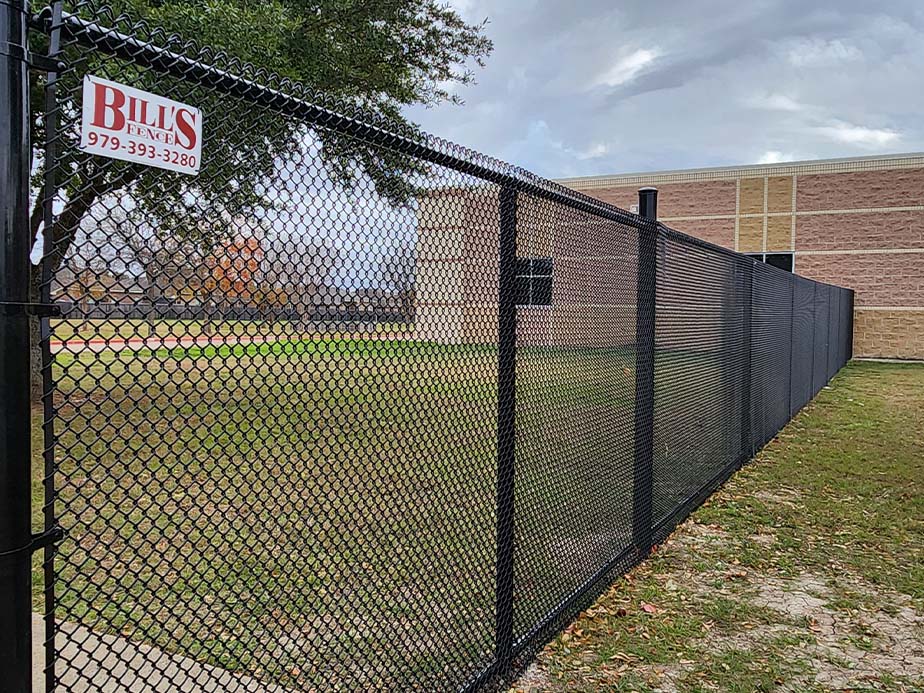 A black chain-link fence runs alongside a grassy area next to a tan brick building. A sign on the fence reads BILLS FENCES with a phone number. Tree branches extend over the fence under a cloudy sky.