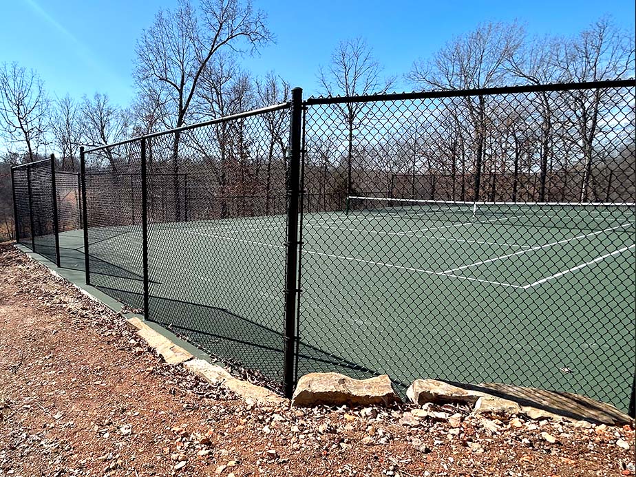 A fenced outdoor tennis court with a green surface sits surrounded by leafless trees and a dirt pathway under a clear blue sky. Rocks line the edge of the court.