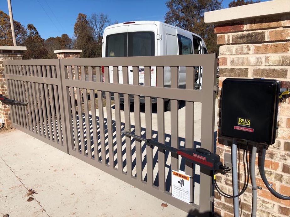 A white van waits behind a closed brown automatic sliding gate, installed between two brick pillars. The gate features warning signs and motor controls mounted on the right pillar. Trees are visible in the background.