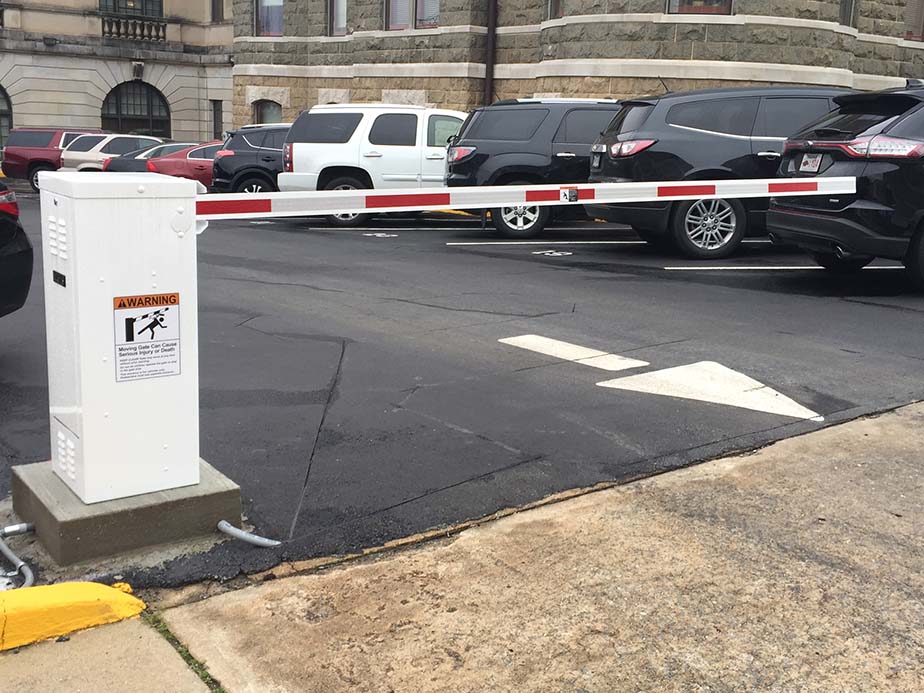 A parking lot entrance with a red and white automatic barrier gate blocking access; several parked vehicles and a beige stone building are visible in the background.