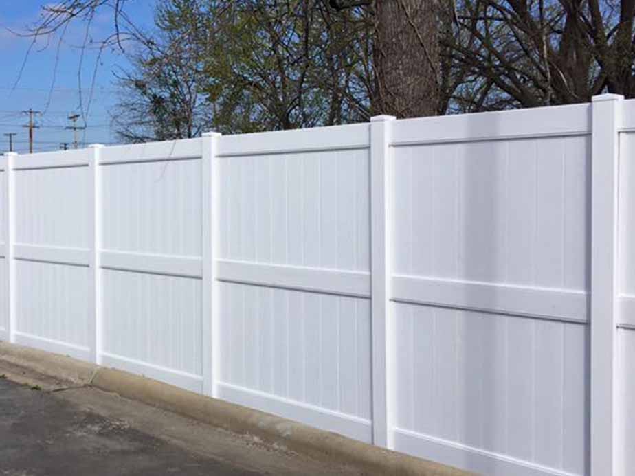 A tall, white vinyl privacy fence with vertical panels stands along a paved area, with trees and blue sky visible in the background.