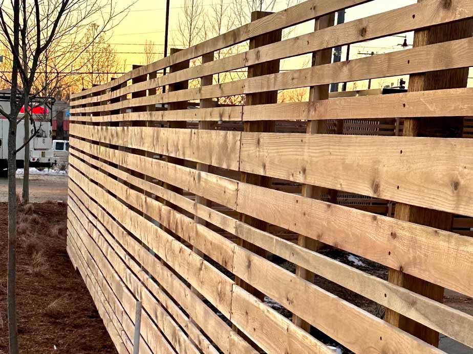 A close-up view of a wooden slat fence runs alongside a sidewalk at sunset, with trees and utility poles visible in the background.