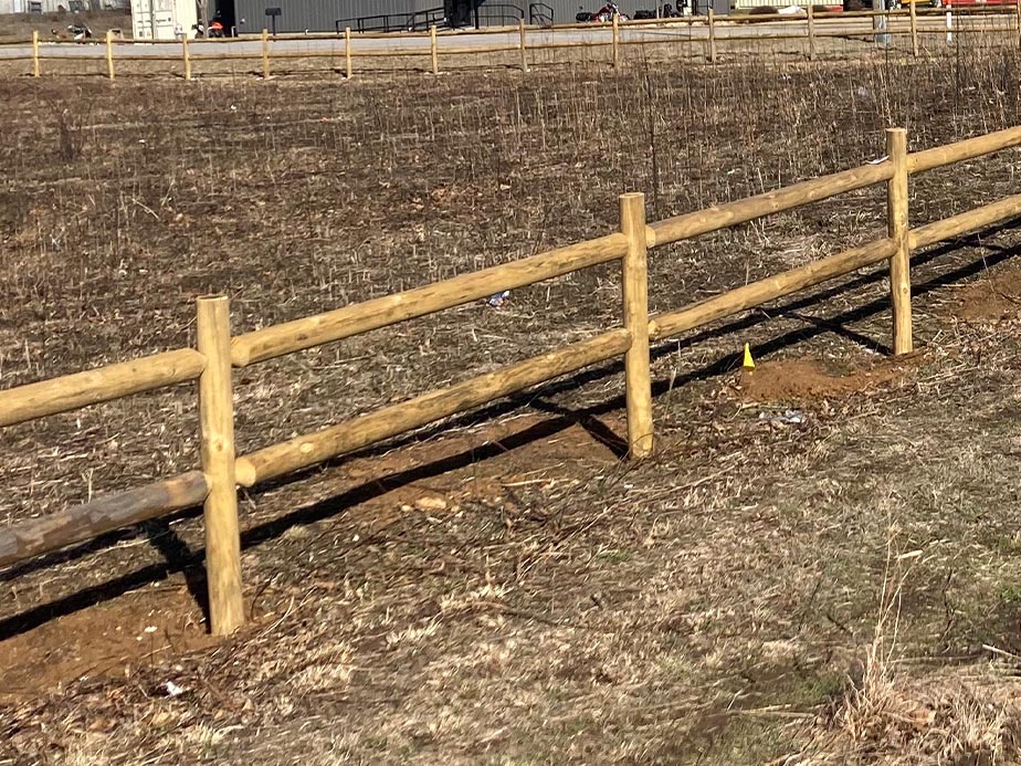 A simple wooden fence runs along a barren, dry field with sparse grass and leafless vegetation. Some buildings are visible in the distant background.