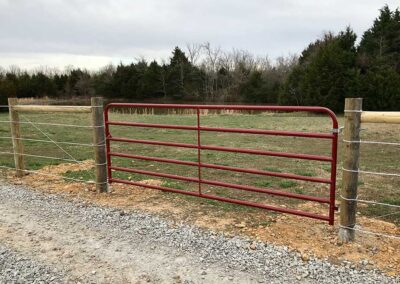 A red metal farm gate is attached to wooden fence posts, with wire fencing on each side. Gravel covers the ground in front, and there are grassy fields and trees in the background under a cloudy sky.
