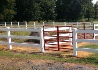 A white wooden fence surrounds a grassy area, with a red metal gate positioned away from the opening in the fence, making it unusable as an entry or exit point. Trees and a dirt path are in the background.