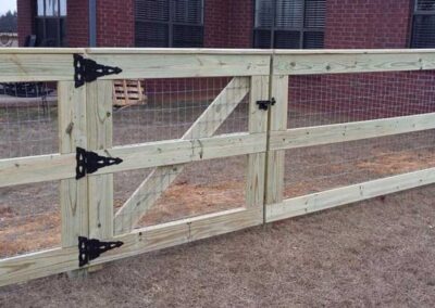 A wooden fence with wire mesh panels and a gate, featuring large black metal hinges and a latch, stands on dry grass in front of a brick house with several windows.
