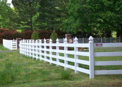 A long white wooden fence lines a grassy area, with trees and shrubs in the background. A sign with red text is attached to one section of the fence. The scene appears to be in a rural or suburban setting.