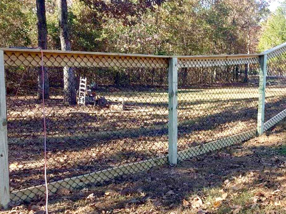 A wooden fence with wire mesh on uneven ground, creating a wavy, non-level appearance. Trees and fallen leaves cover the area beyond the fence, with a couple of chairs visible in the background.