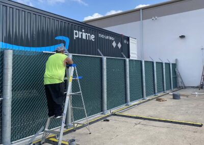 A worker in a neon yellow shirt stands on a ladder installing privacy mesh on a chain-link fence beside a large Amazon Prime trailer in an industrial area, with tools and materials on the ground.
