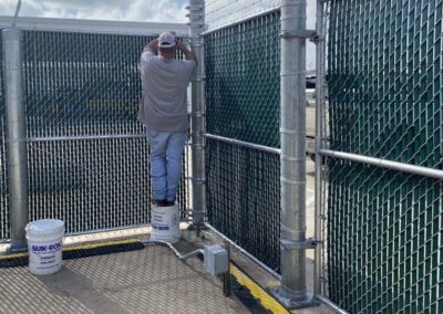 A man stands on two stacked buckets labeled QUIK-ROK to reach the top of a chain-link fence, working with tools near the barbed wire under a partly cloudy sky.