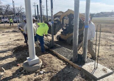 Construction workers pour and level concrete into a rectangular frame at a worksite, using a small skid-steer loader. Metal poles and construction materials are visible around the site.
