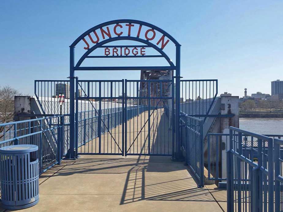 A gated blue entrance to the Junction Bridge pedestrian walkway, with Junction Bridge written overhead. The bridge extends over a river towards a city skyline in the background under a clear sky.