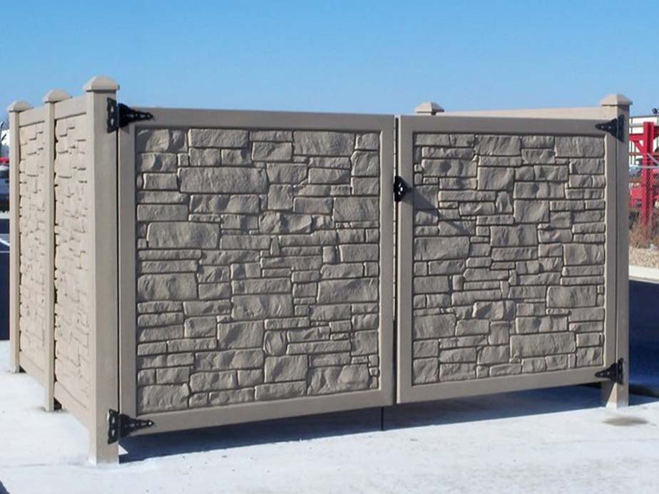 A tan and gray privacy fence with a stone-like textured pattern, metal hinges, and posts, set on a concrete slab under a clear blue sky.