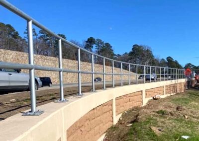 A metal railing runs along a raised sidewalk beside a road, with cars driving past. Trees and a stone wall line the opposite side under a clear blue sky.