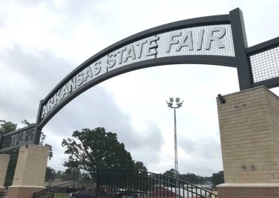 A large metal archway sign reads ARKANSAS STATE FAIR in bold white letters, supported by brick columns, with trees and a cloudy sky in the background.