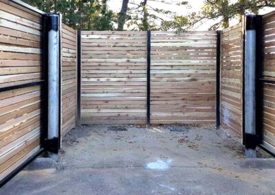 A three-sided enclosure made of horizontal wooden slats and black metal supports stands on a concrete slab, surrounded by trees and outdoor greenery.