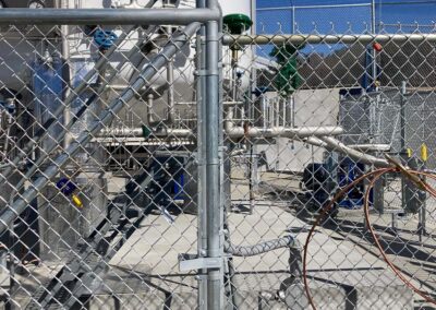 A fenced industrial area with metal pipes, tanks, and equipment visible behind a chain-link gate under a sunny sky. Various cables and mechanical components are present.