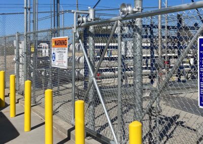 A chain-link fence with warning signs and bright yellow bollards surrounds an industrial facility with electrical equipment and power lines visible behind the fence under a clear blue sky.