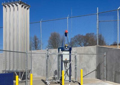 A fenced industrial area with a tall silver gas cylinder rack, control equipment, yellow safety bollards, and a concrete wall topped with chain-link fencing under a clear blue sky.