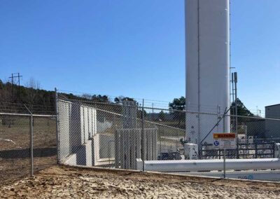 Chain link fence surrounds an industrial facility with a tall white storage tank, warning signs, and piping; trees and a clear blue sky are in the background.